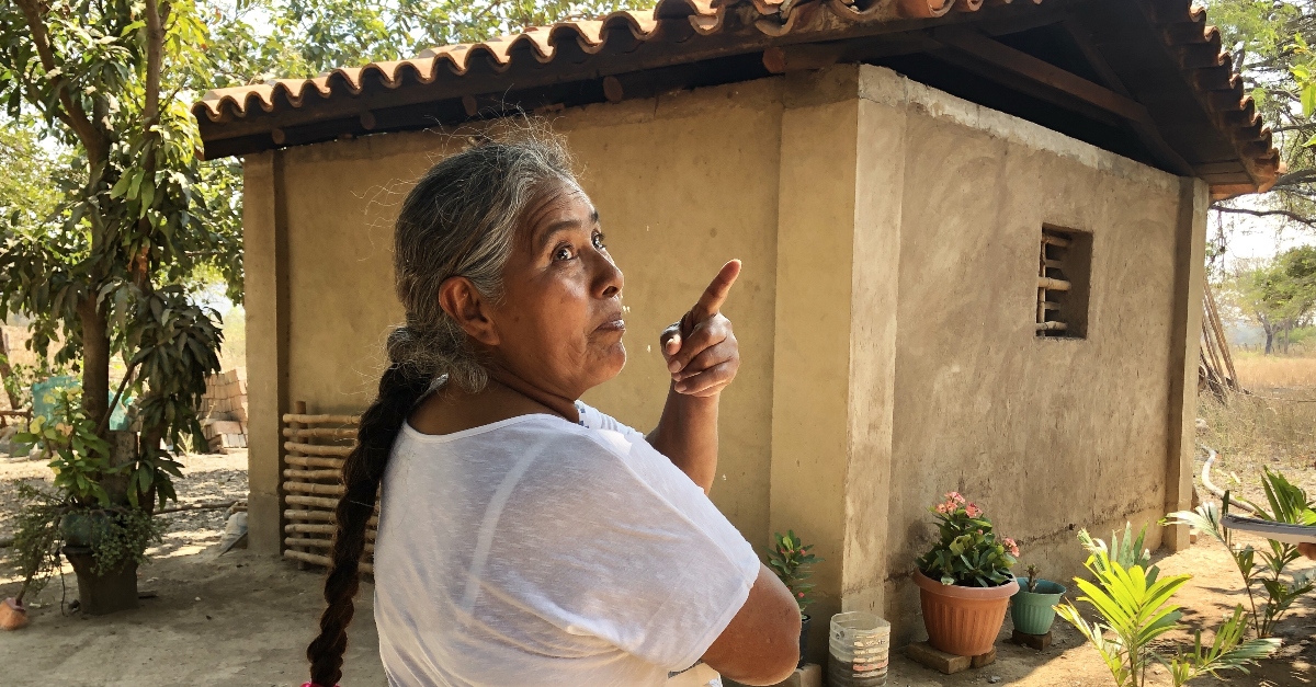 A woman stands with her back facing the camera and in front of a mud brick house. She is looking over her shoulder to the right while pointing with her left hand.