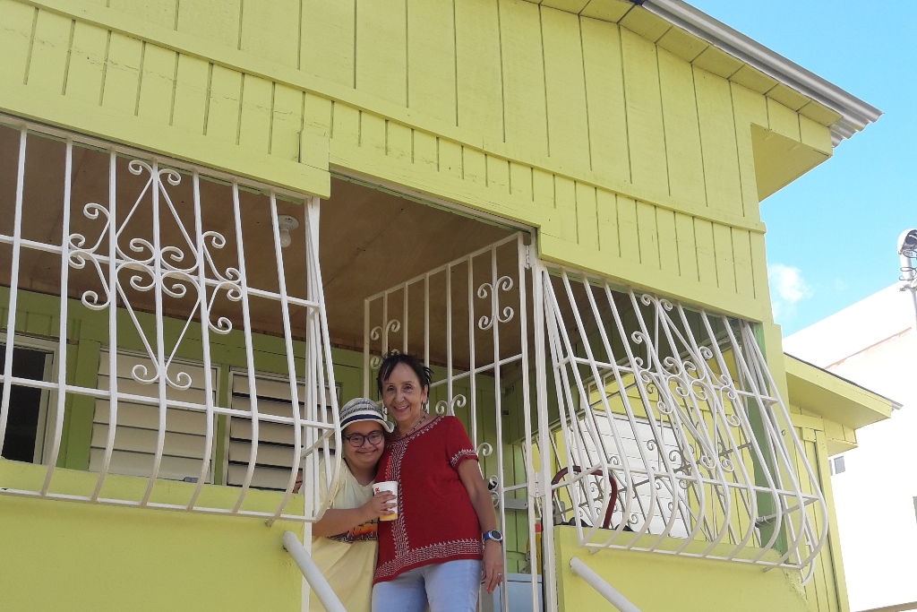 A woman and a young girl stand in the doorway of a yellow house with white wrought iron over the windows and door.