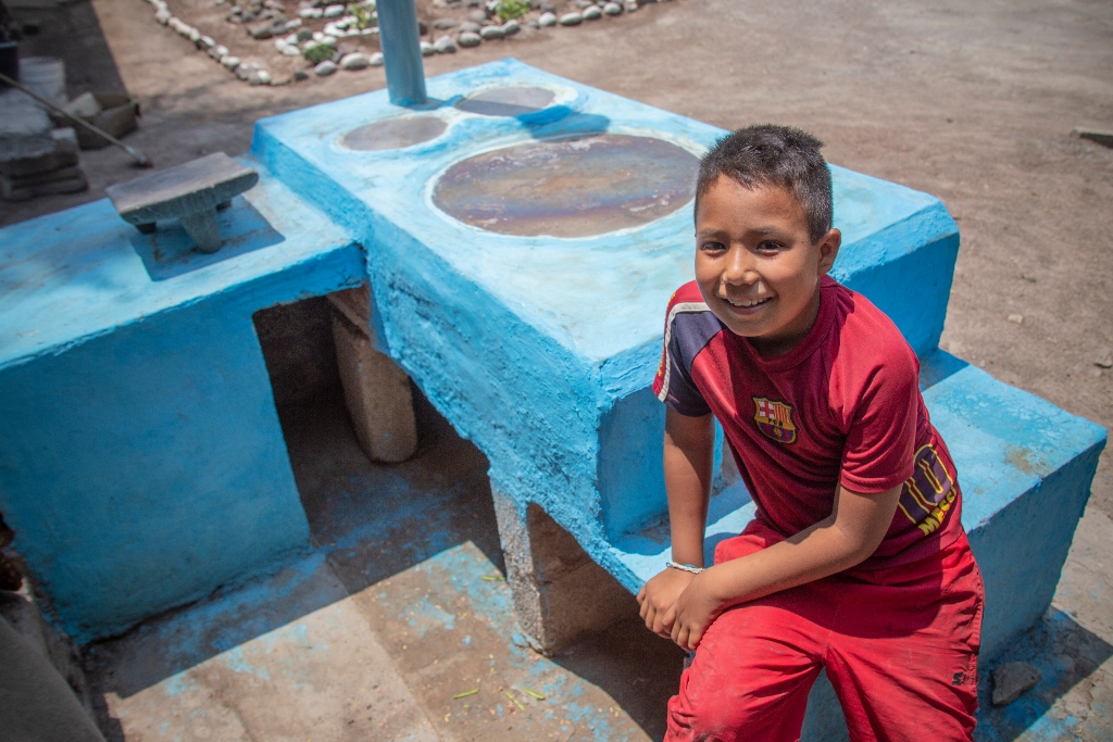 A young boy in a red shirt and pants sits on the side of a blue stove made from mortar 
