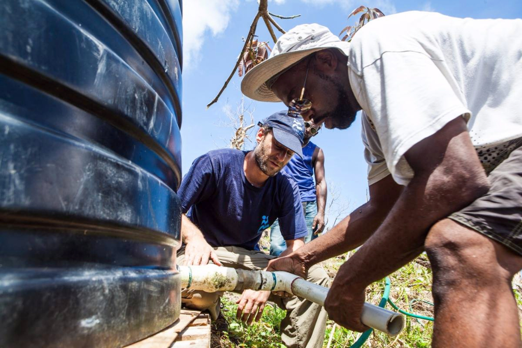 A man in a hat, glasses, and white t-shirt adjusts a pipe connected to a black water tank. A man in a blue shirt and hat helps him to the side.
