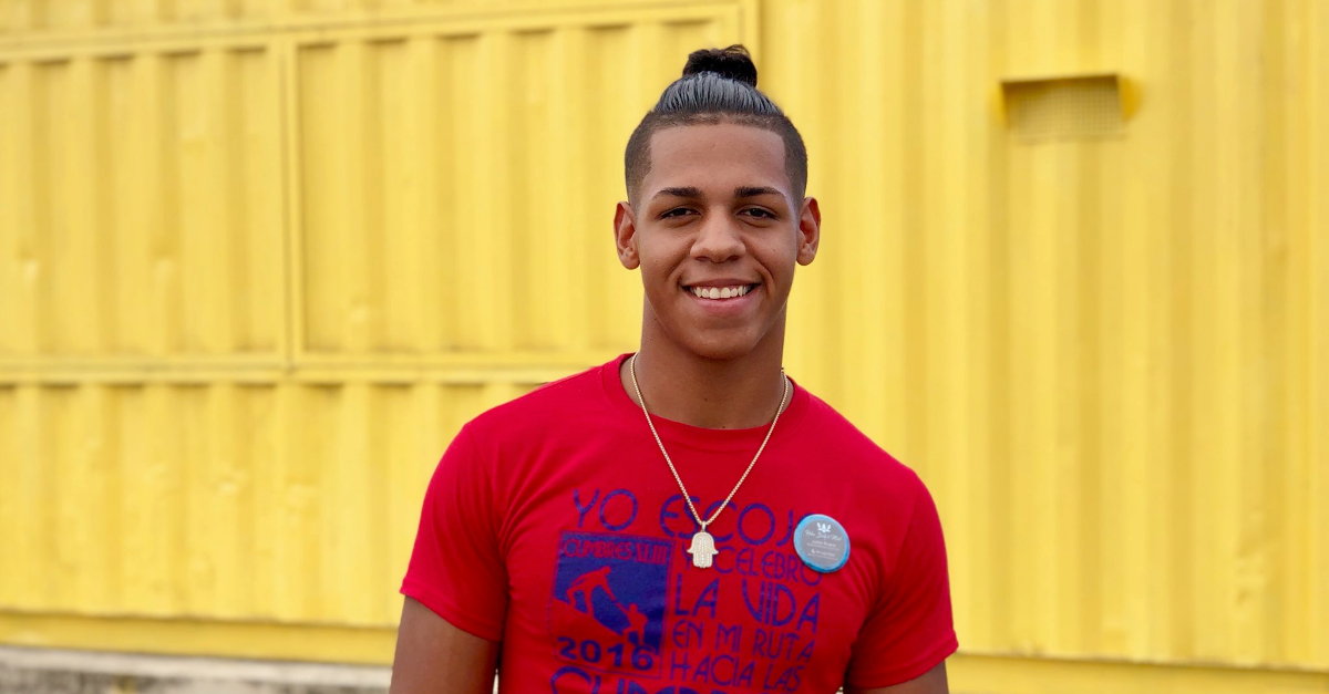 A teen boy in a red t-shirt and necklace stands in front of a bright yellow shipping container