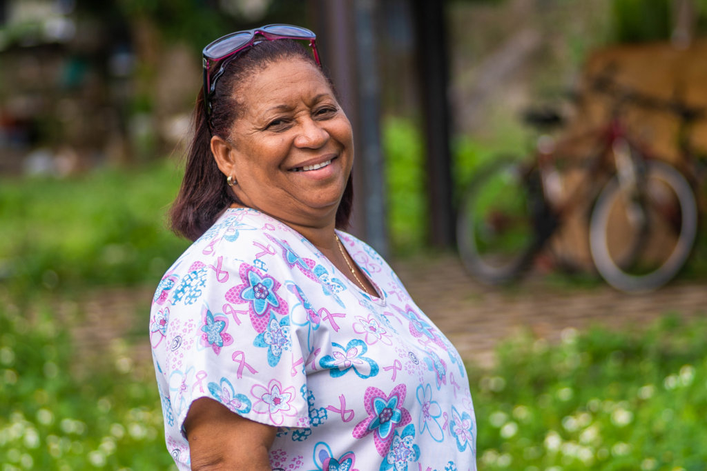 A woman in a flower-patterned shirt and glasses on her head smiles. A bike and green grass with flowers are out of focus in the background. 