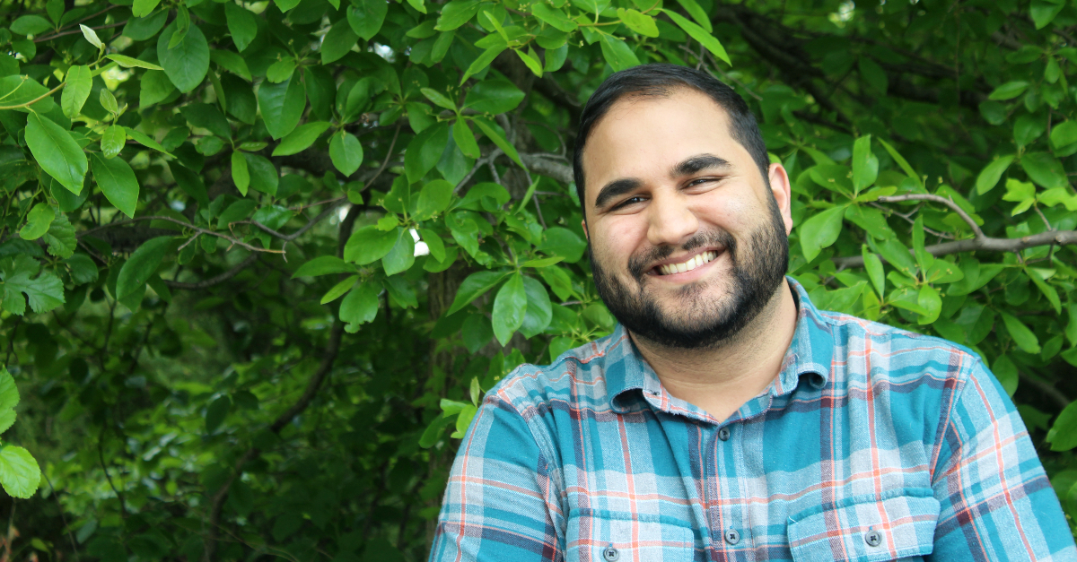 A man in a blue plaid shirt stands smiling in front of a green tree. He sends international donations