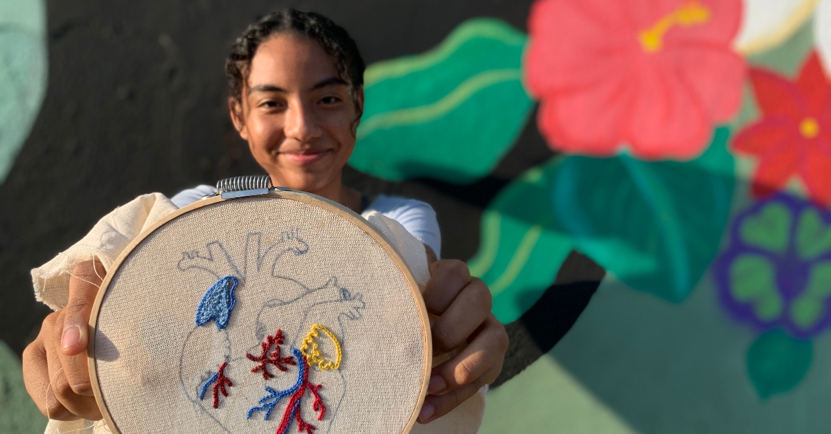 A young smiling girl holds fabric with cross-stiching in the shape of an anatomical heart in the foreground.