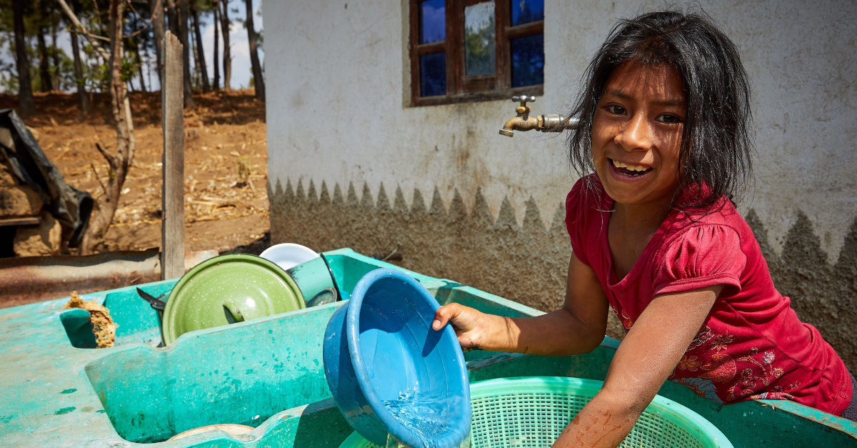 A young girl holds a blue bowl while standing in front of a sink. A house and a water faucet are in the background.