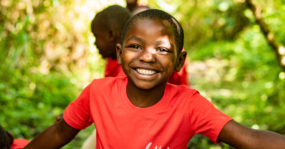 A boy in a red shirt smiles as he holds his arms out to the sides. Other children and trees are out of focus in the background.
