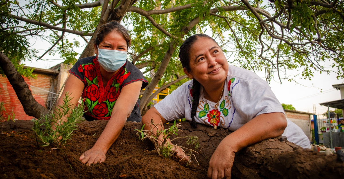 Two women work with their hands in the soil. One is wearing a mask, and the other is looking up and to the side without a mask on.