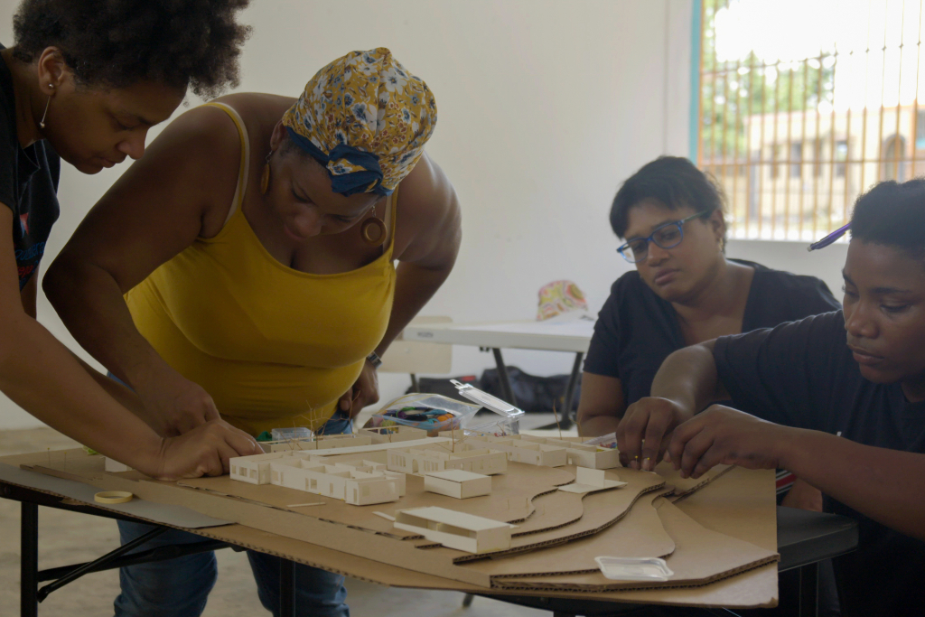A group of people sit and stand around a table where model buildings made out of wood sit on top of cardboard.