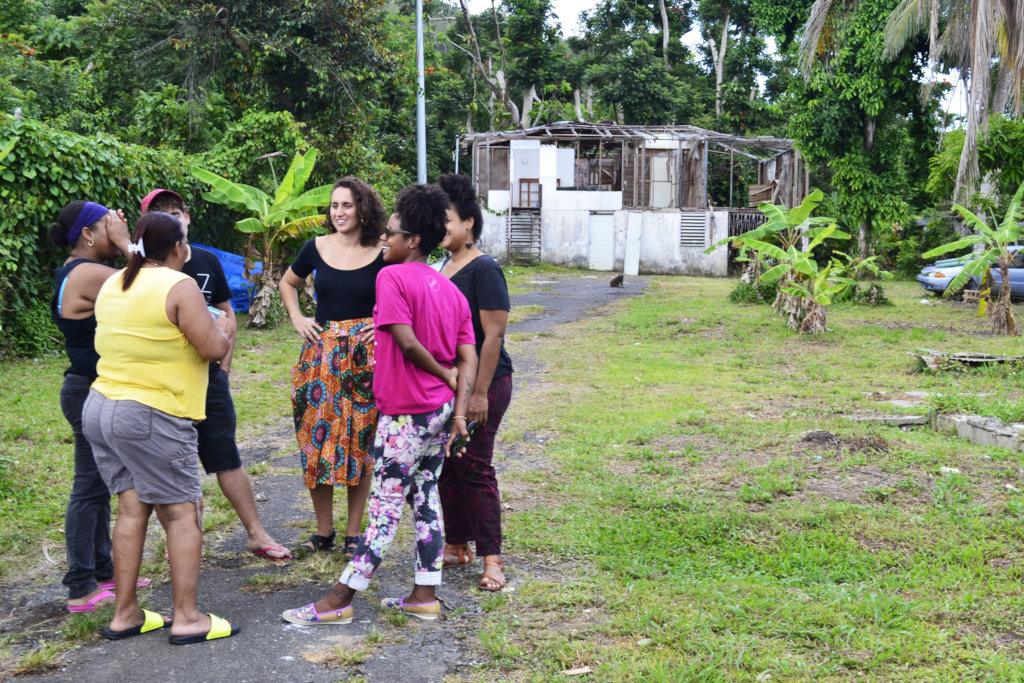 A group of women stand on a gravel path. A white house destroyed by Hurricane Maria is behind them. 