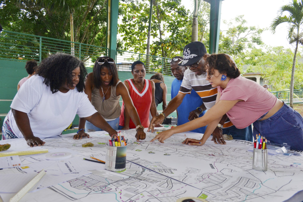 A group of people stand around a table with blueprints spread out in front of them and point to different sections.