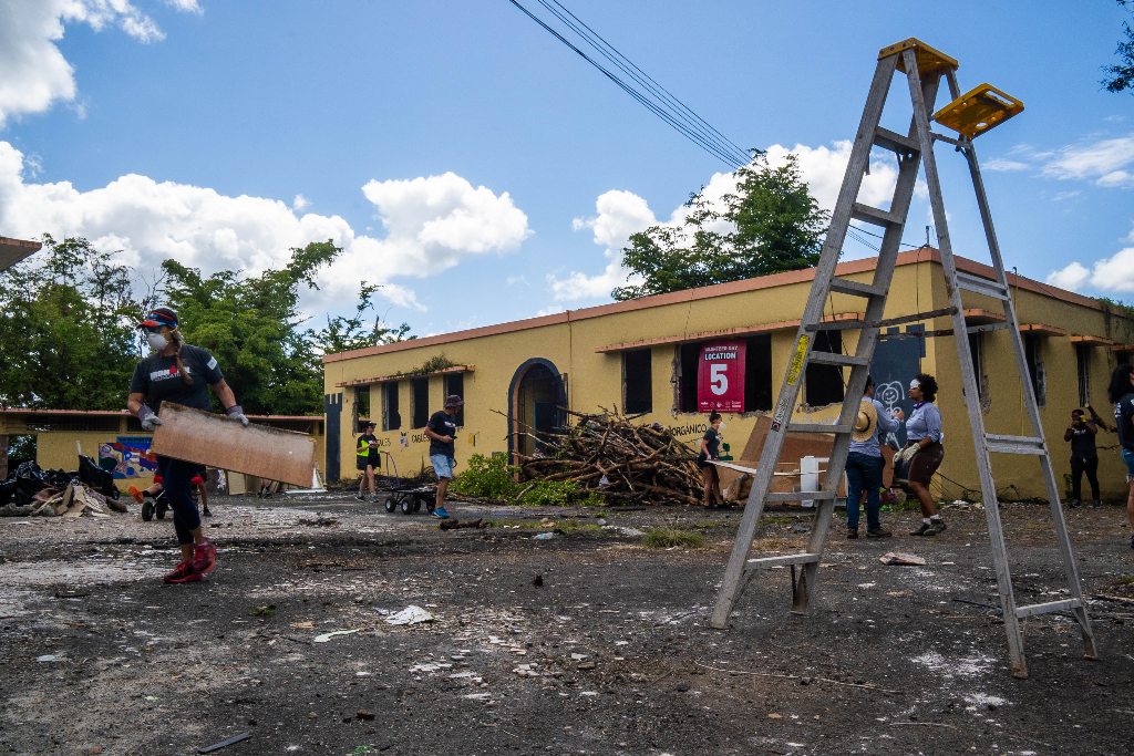 People move debris surrounding a yellow building. A ladder is in the foreground in the right corner.