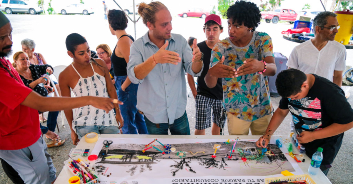 Two people look at each other and gesture with their hands to illustrate the slope of a pointed roof. Other people stand around them, and on the table in front of them are paints and a piece of paper with a black and white image.