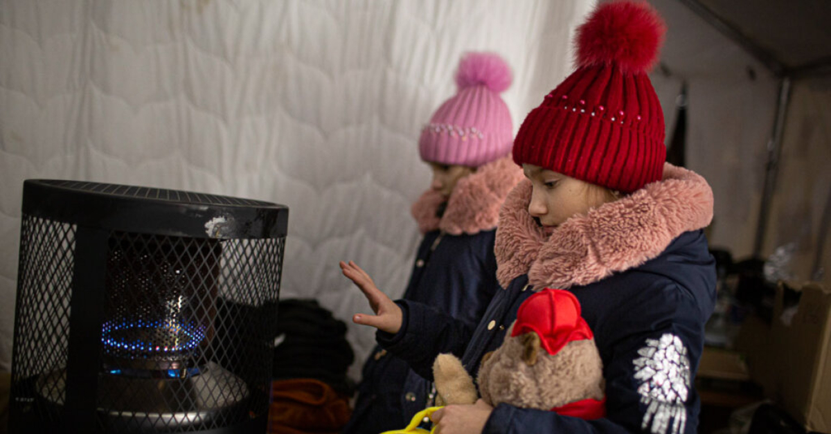 Two young girls wearing hats and jackets warm themselves near a heater. One girl holds stuffed animals.