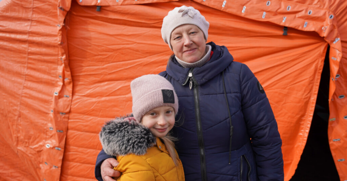 A woman and her daughter wear winter jackets and hats in front of an orange tent. The mother's arm is around her daughter.
