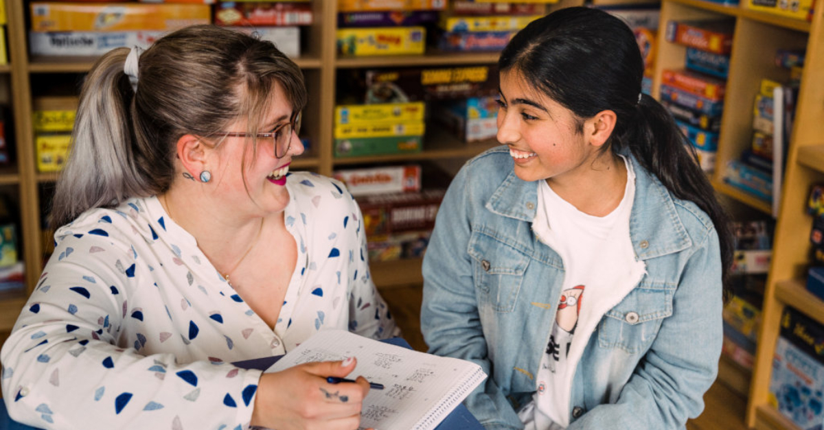 A person wearing a white patterned shirt and glasses holds a pen above a notebook while talking to a young student wearing a jean jacket. Games are on a shelf in the background behind them.