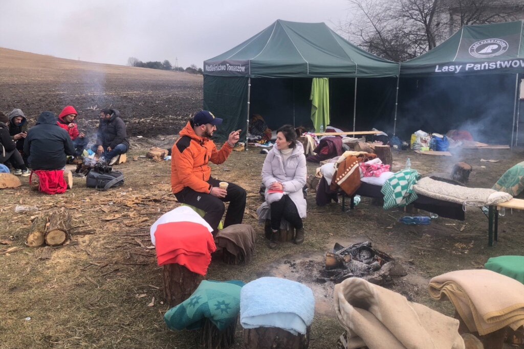 Two people sit on a bench near a dying fire at a refugee camp. Blankets cover benches and logs around them, and other refugees are grouped together in the background.