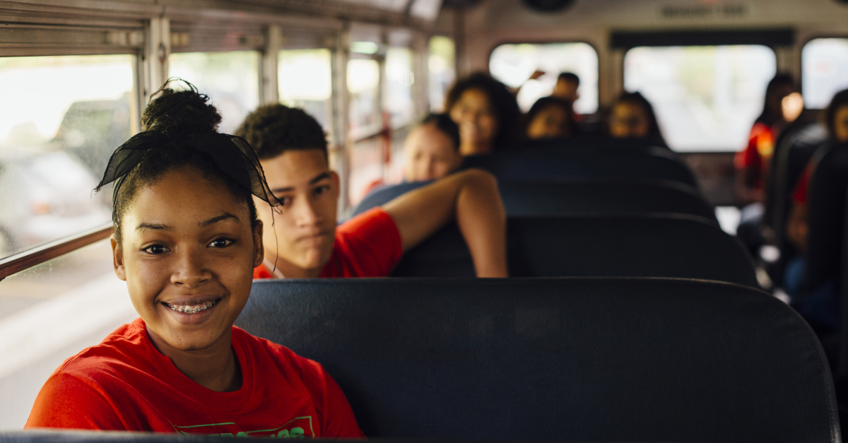 A young student with braces and a black ribbon tied in their hair smiles in the seat of a school bus. Other students are sitting in seats in the background