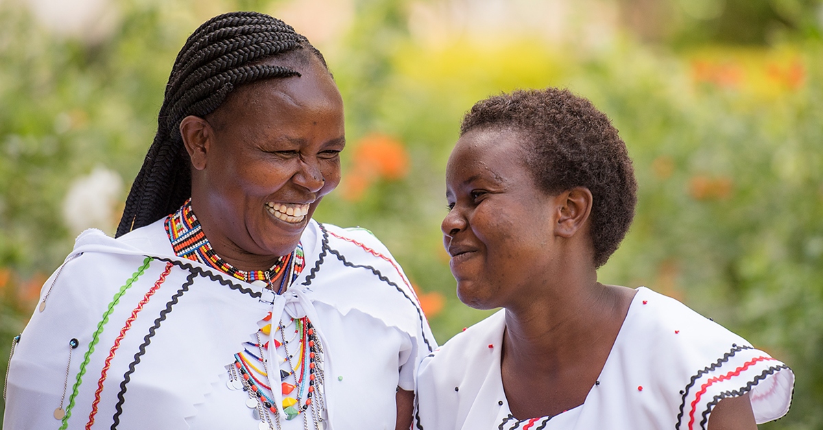 Two women in white blouses standing facing each other and laughing. Flowers and grass are out of focus in the background