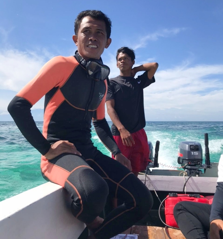 A man in a black and orange wetsuit sits on the edge of a boat. Blue water and a clear blue sky are in the background