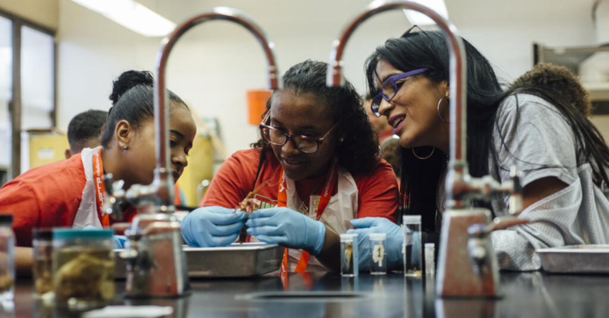 Two students wearing blue disposable gloves work with a woman in a science lab. Vetting nonprofit partners