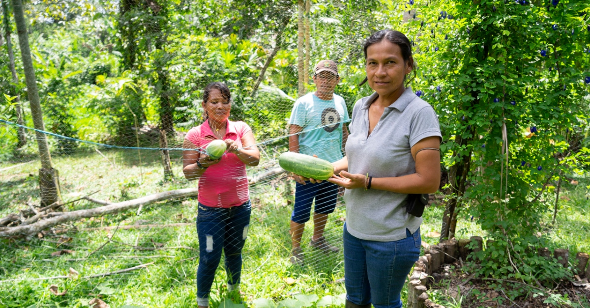 A woman holds a cucumber in her hands and is surrounded by trees. Two people stand behind a net in the background