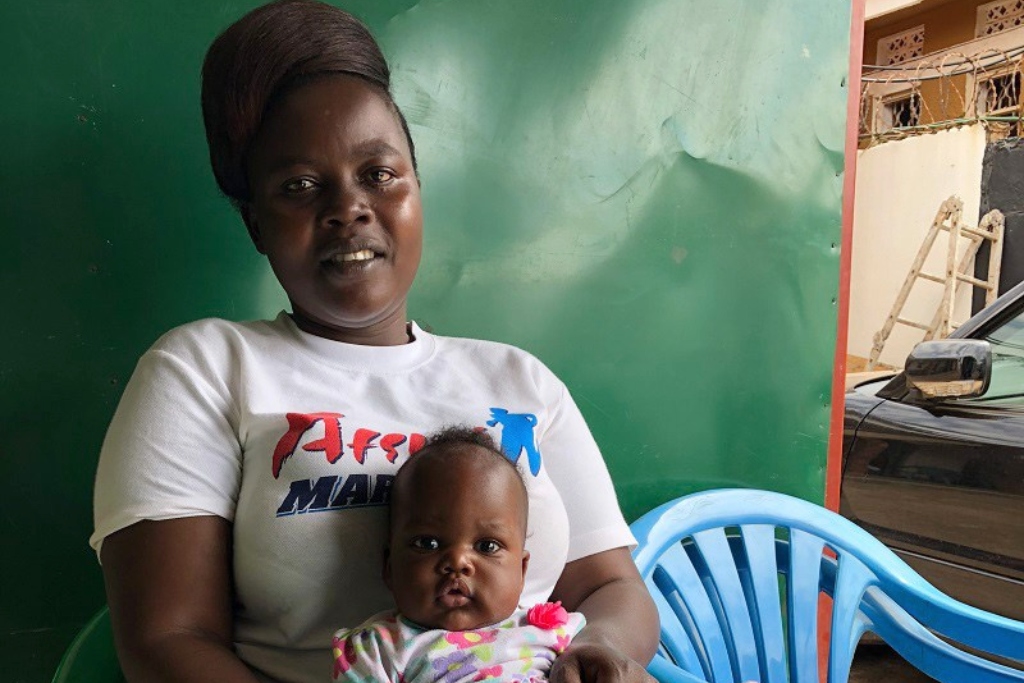 A woman in a white t-shirt holds her baby in her lap. A green wall is behind her, and blue plastic chairs are at her side