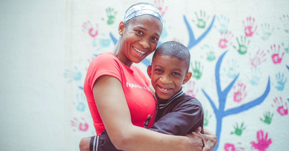 A mother and a son smile and embrace. A mural of tree branches and handprints covers the wall behind them.