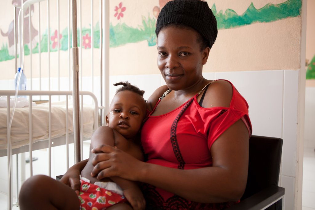 A mom in a red shirt holds here baby on her lap near a hospital bed. Flowers and green grass are painted on the wall behind them. 