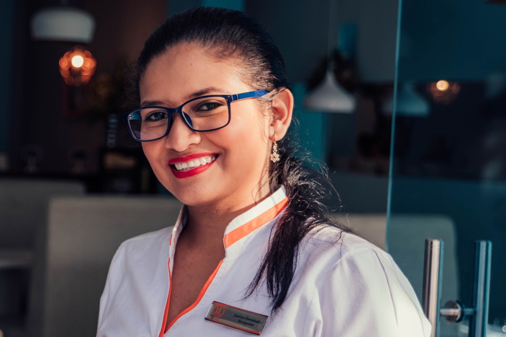 A woman in a white uniform and blue glasses smiles. Hanging lights and booths are out of focus in the background