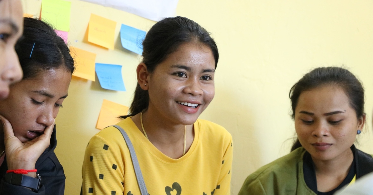 A young woman wearing a yellow smiles and looks off to the side. Other young women sit next to her, and there are colored sticky notes on a pale yellow wall in the background