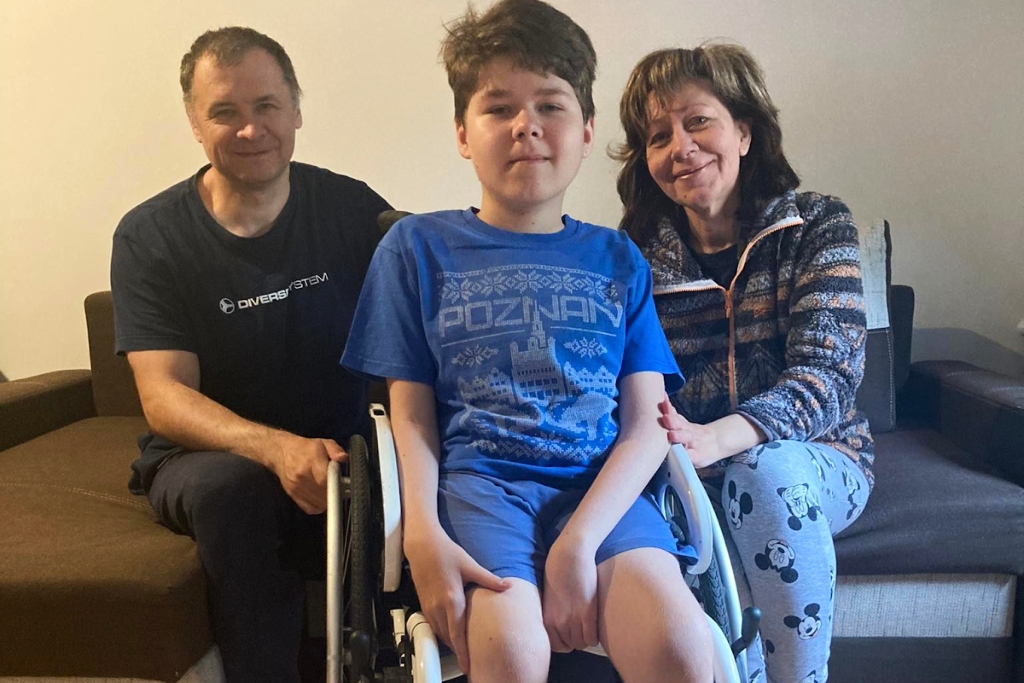 A young boy in a blue shirt and shorts sits in a wheelchair. His parents sit behind him on either side, smiling