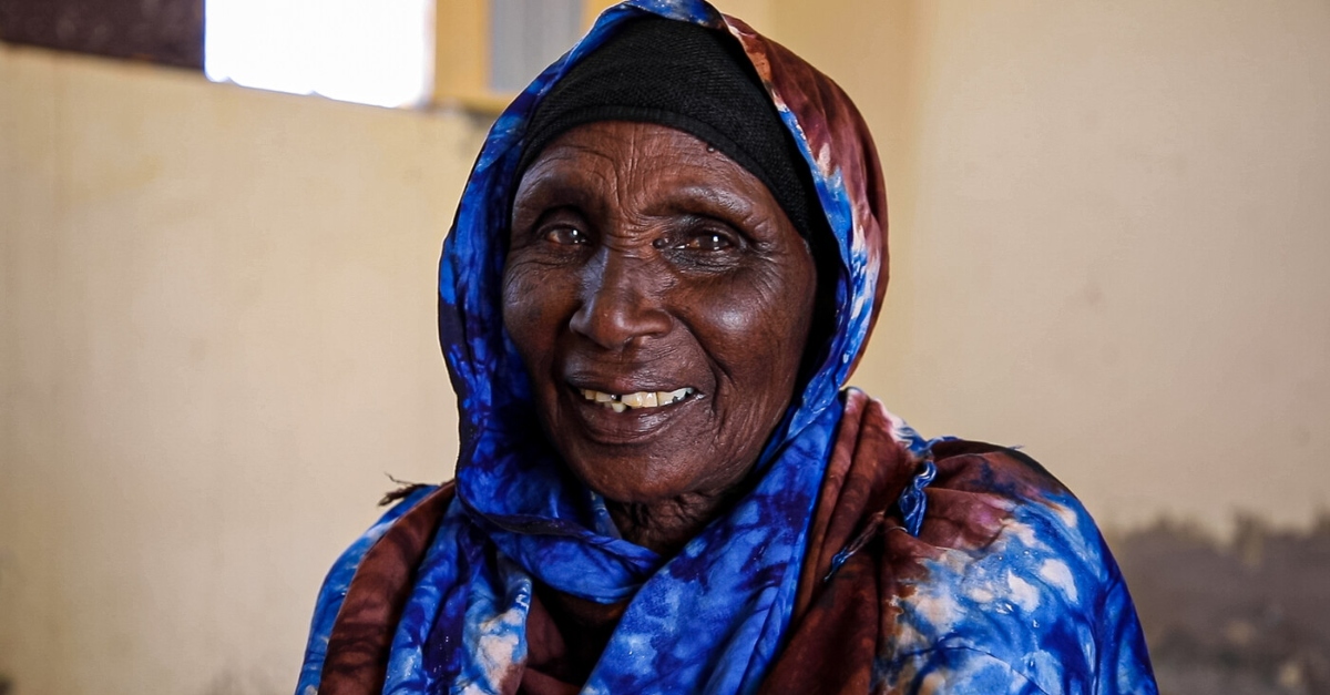 A woman in a tie-dyed hijab smiles. A pale yellow wall is out of focus behind her. Equity in crisis response