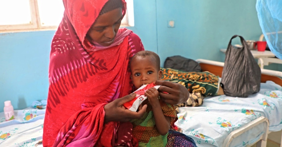 A mother in a red hijab sits on a hospital bed and holds her child while feeding them a nutritious peanut paste.
