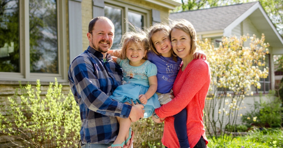 A man wearing blue and women wearing red hold two young daughters in front of a home