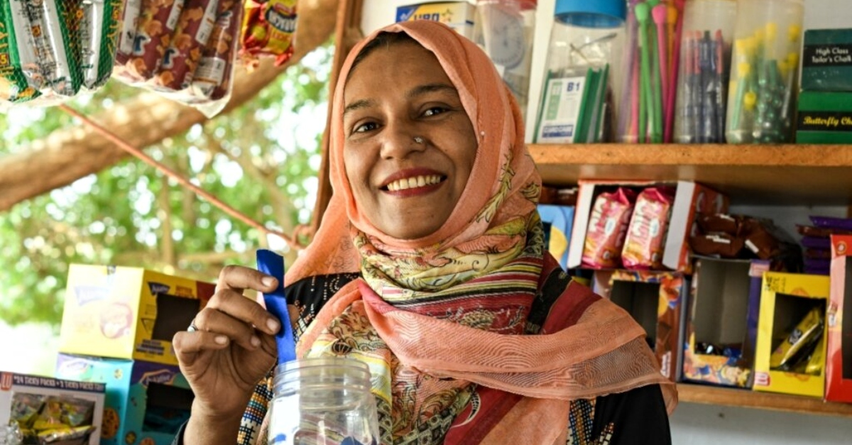 A woman wearing a peach colored headwrap smiles as she pulls a ribbon out of a jar. Shelves stacked with goods are in the background