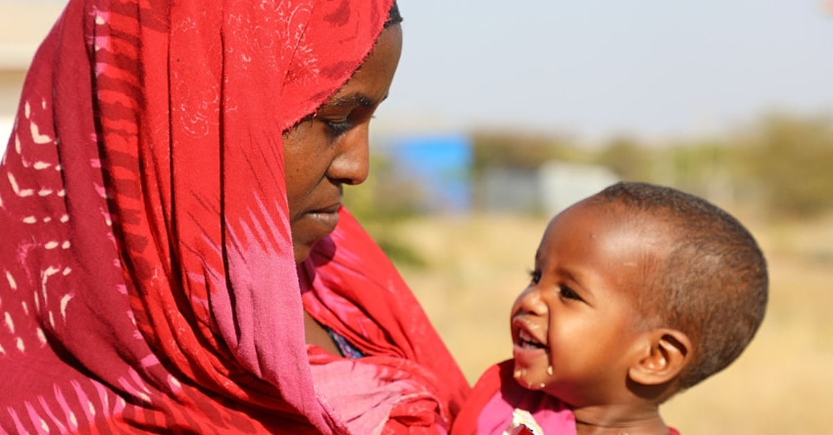 A mother in a red hijab holds her child who smiles at her.