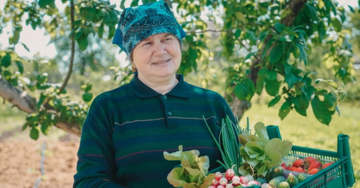 A woman in a green and blue shirt and a blue patterned head scarf stands holding a case of vegetables. A tree is in the background