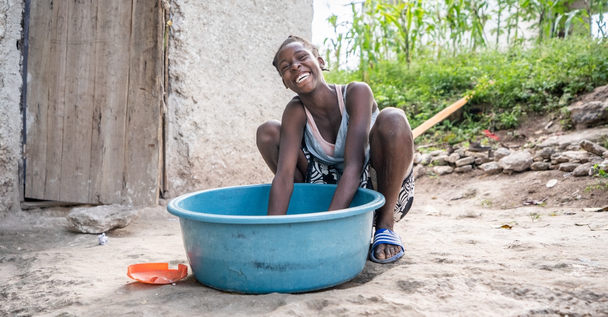 A smiling woman sits behind a big plastic bucket and has her hands inside. A wooden door and concrete building are in the background