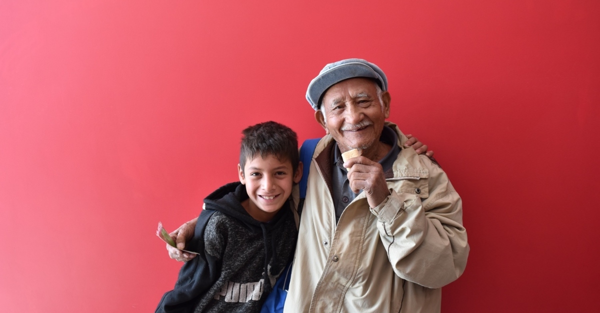 An elderly man and a young boy smile and embrace each other in front of a red wall