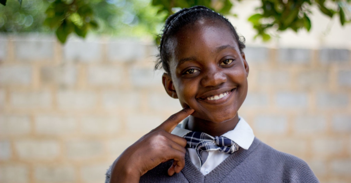 A young student wearing a gray and white uniform smiles and holds her index finger next to her cheek. Leaves are out of focus in the background. donate from your IRA