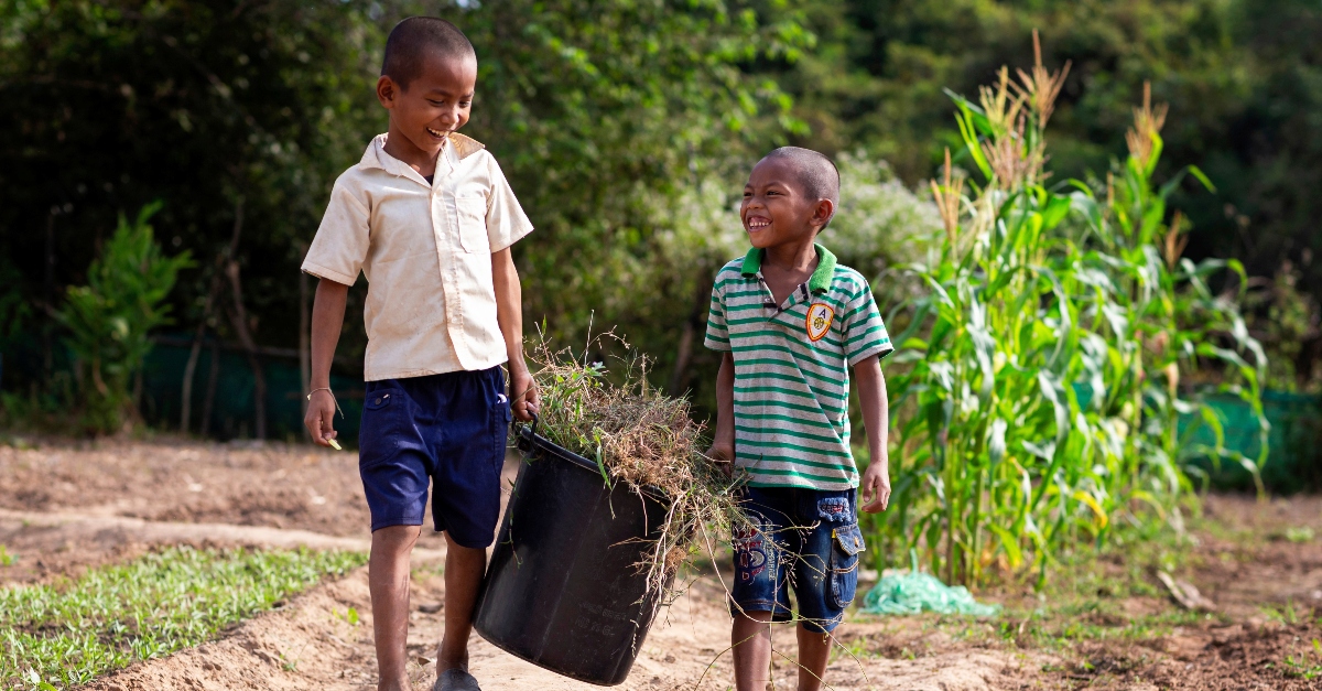 Two young kids smile while each holds a black bucket filled with grass and weeds on one side. Community led