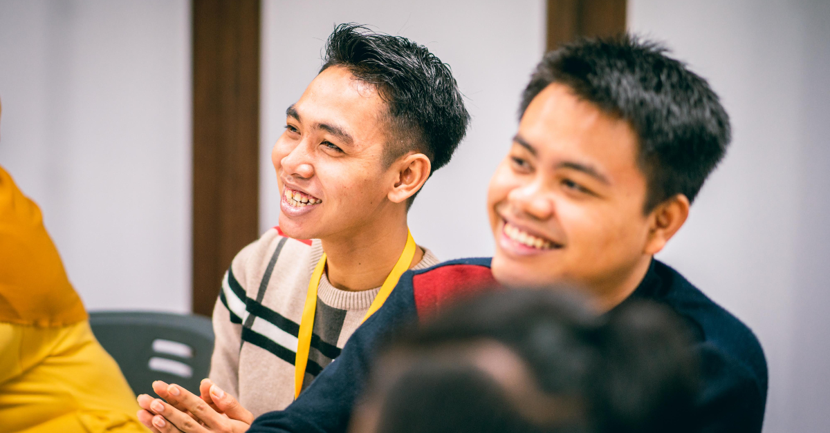 Two young men in a larger group, watching off to the left and smiling.