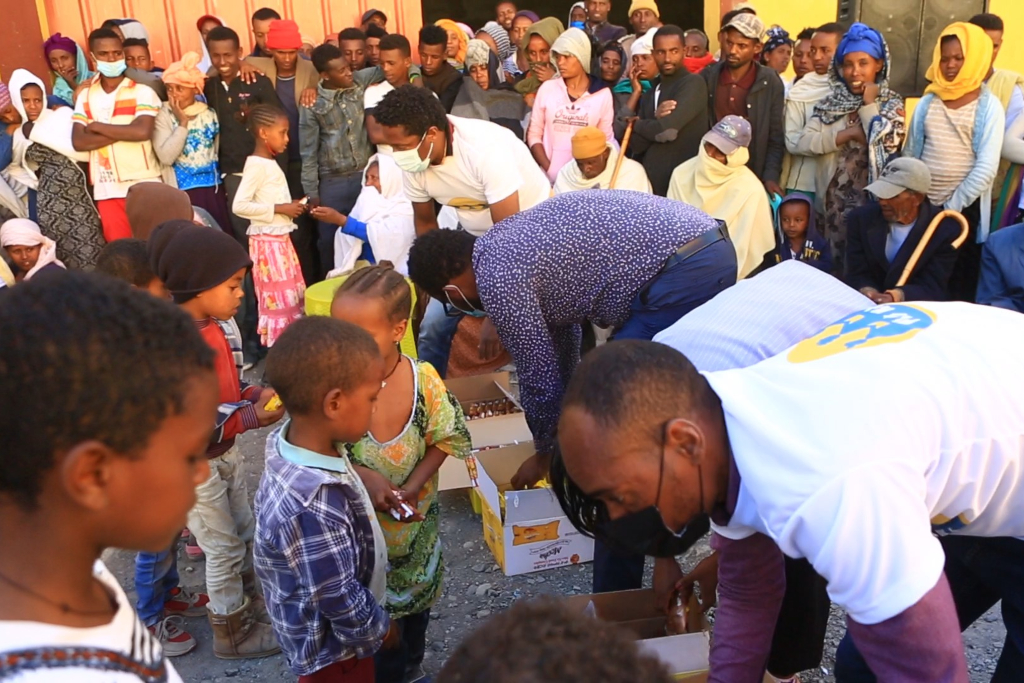 a crowd gathers around boxes filled with food, distributed by men wearing masks responding to crisis