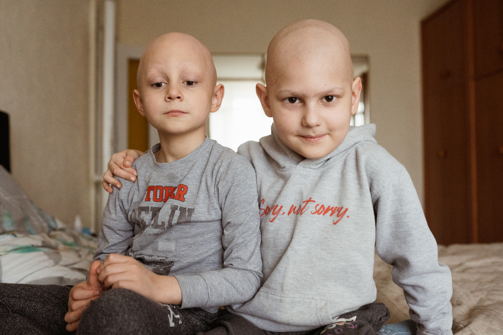 Two children in cancer ward sit together one's hand around the other's shoulder, looking at the camera