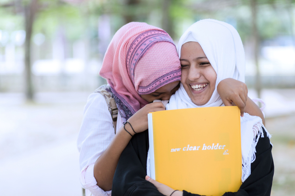 Two girls giggle together, holding school books - photos of 2022
