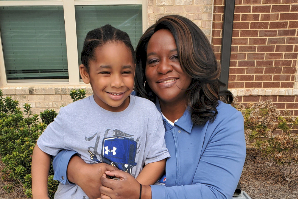 A woman with her arms around her young son sits in front of a brick building with a window