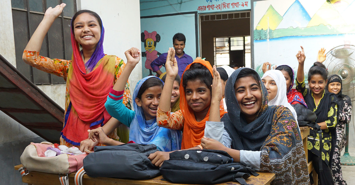 group of young women sit at desks raising their hands and smiling and laughing. community voices