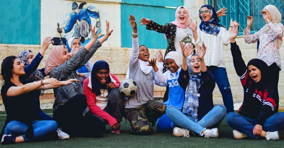 Girls with their hands in the air smile and shout on a soccer field during a program to welcome refugees. A soccer ball bounces between them.