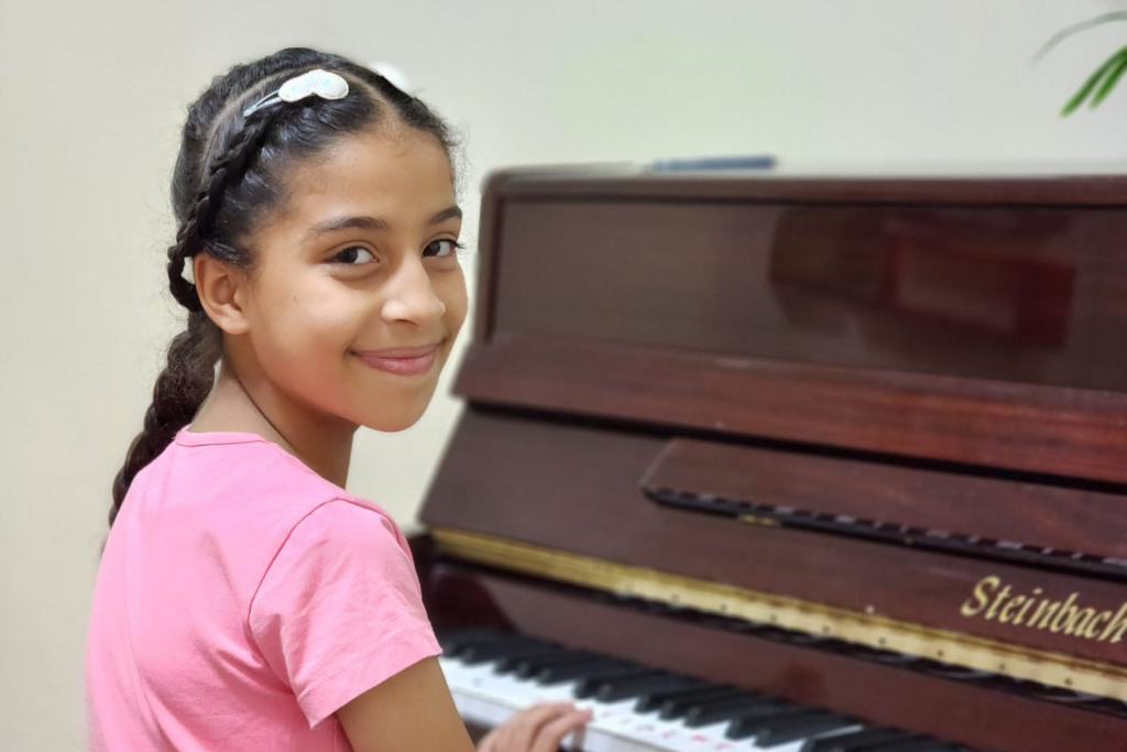 girl sits at piano and smiles over her shoulder at the camera refugee notes