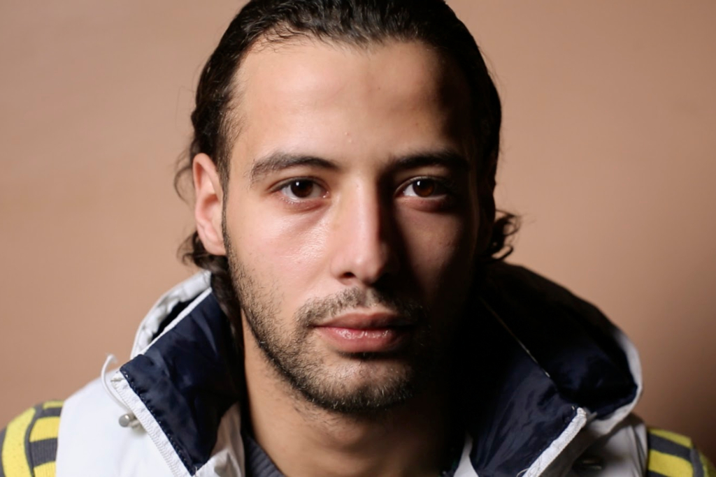 man wearing windbreaker looks solemnly at the camera with his hair tied back